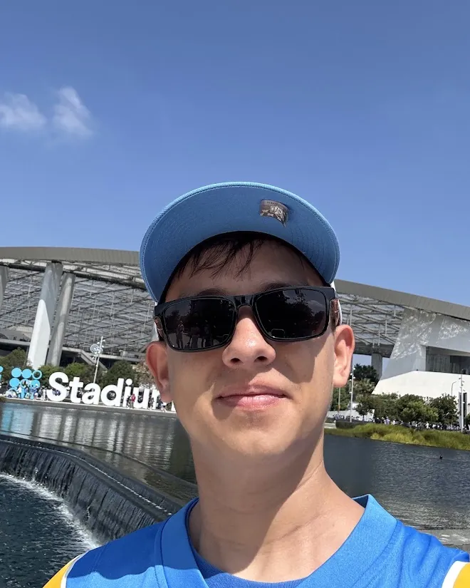 Victor's selfie in a cap and dark sunglasses in front of SoFi Stadium on a clear day.